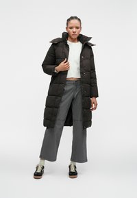 Garšs tumši brūns pūkains mētelis ar augstu apkakli, quilting dizainu un pogu priekšpusi. Kombinēts ar pelēkajām platām biksēm un melnām kedām.