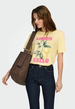 Kobieta w żółtej koszulce z grafiką cytryny i ciemnych dżinsach, niosąca dużą brązową torebkę na ramię, lekko uśmiechająca się i patrząca w dół.