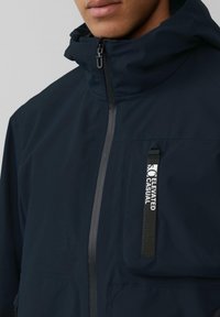 Marynarka w kolorze granatowym z wysokim kołnierzem, pełnym zamkiem, kieszenią na klatce piersiowej z detalem logo oraz kontrastującym wykończeniem zamka. Gładki, lekki materiał.