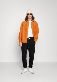 Camisa de botones naranja sobre una camiseta blanca, pantalones sudadera negros con logo y zapatillas blancas. Los accesorios incluyen un gorro crema y gafas de sol.
