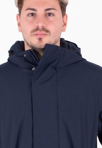 Giacca impermeabile blu navy con colletto alto e cappuccio. Presenta un design minimalista, una leggera lucentezza e una chiusura frontale con zip e bottoni a pressione.