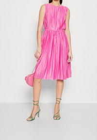 Różowa sukienka plisowana z okrągłym dekoltem i tasiemką w talii. Połączona z zielonymi sandałami na obcasie i małą okrągłą różową torebką. Gładka faktura.