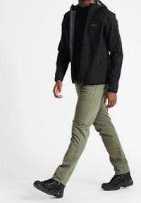 Giacca softshell nera con interno grigio, cappuccio e tasche con zip; pantaloni verde oliva; stivali robusti neri con lacci e suole antiscivolo.
