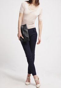 Vaaleanbeige istuva yläosa, jossa on pyöreä kaula-aukko, tummansiniset räätälöidyt housut ja off-white nilkkaketjupohjalliset kengät, ja hän pitää mustaa kierrätettyä clutch-laukkua.