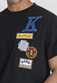 Černé bavlněné tričko s různými vyšívanými nášivkami: velké modré "K", žluté "Gianni Kavanagh", šedé logo a červené kruhové logo.