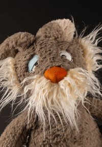 Jouet en peluche marron avec une barbe blanche ébouriffée, un nez orange, des oreilles tombantes et des yeux cartoon dépareillés sur un fond noir.