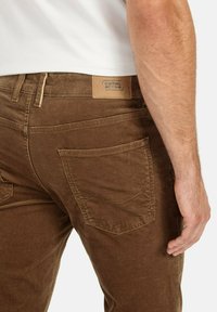 Un pantalon en velours côtelé marron avec une coupe droite, des poches arrière cousues et un patch en cuir beige sur la taille.