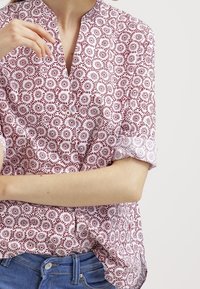 Blouse à manches courtes blanche et rouge, présentant un motif floral, un col en V et des poignets retroussés. Confectionnée en tissu léger.