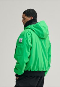 Grüne Kapuzenjacke aus glattem Stoff, mit einem schwarzen Kragen, elastischen Bündchen, einer Vorderreißverschlusstasche und einem Logo-Patch auf dem Ärmel.