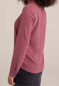 Chemise à manches longues en tissu mauve texturé, présentant des motifs floraux en relief et des poignets boutonnés, conçue avec une coupe décontractée et un ourlet arrondi.