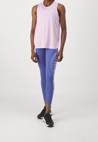 Person som bär en ljusrosa ärmlös topp, blå Under Armour leggings med logotyp på sidan, och svarta löparskor som står mot en enfärgad bakgrund.