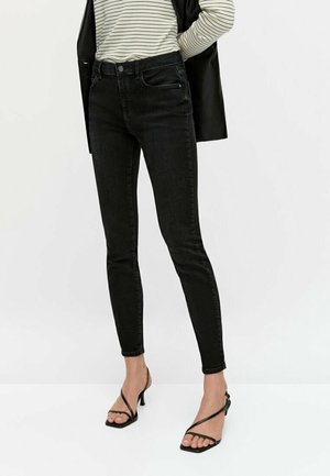 Zwarte slim fit jeans gecombineerd met zwarte sandaalhakken met bandjes en een gestreept shirt met lange mouwen, met de handen achter de rug gevouwen.