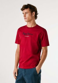 Raudona medvilninė marškinė su trumpomis rankovėmis, apvaliu iškirpimu ir tamsiai mėlynu prekinio ženklo logo tekstu "Pepe Jeans London" per krūtinę.