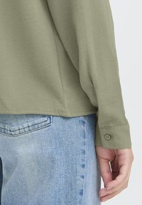 Main droite reposant près de la poche d'un jean bleu clair, portant une chemise à manches longues vert olive avec un poignet boutonné, sur fond uni.