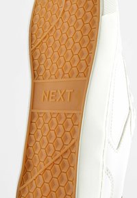 Witte sneakersole met een hexagonaal patroon en een oranje rubberen buitenzool. Het woord "NEXT" is in het midden geperst.