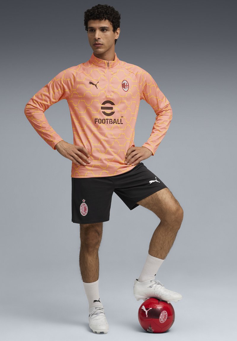 Ružový dlhý rukáv futbalový tréningový top so žltým geometrickým vzorom, čierne kraťasy, biele ponožky a strieborné kopačky; červená lopta pri nohe.