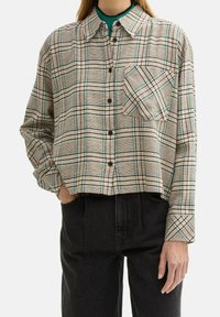Camicia corta a quadri beige con linee nere e teal. Presenta una chiusura con bottoni sul davanti, maniche lunghe e una tasca singola sul petto.