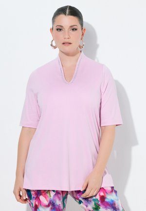 Kvinde med mørkt hår trukket tilbage iført en lyserød, kortærmet top og farverige blomstrede bukser, stående mod en hvid baggrund.