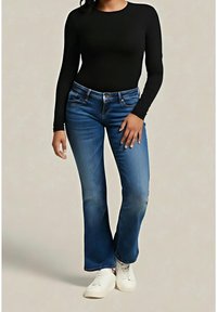 Femme portant un haut noir à manches longues, un jean bootcut bleu et des baskets blanches, debout devant un fond neutre uni.