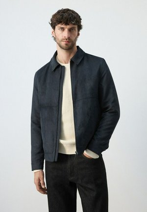 Hombre con chaqueta azul marino con cremallera sobre suéter de punto blanco y pantalones negros, de pie contra un fondo claro y liso.