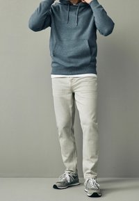 Felpa grigia con cappuccio e tasca a marsupio, abbinata a jeans beige chiaro e scarpe da ginnastica grigie. Outfit casual con tonalità neutre.