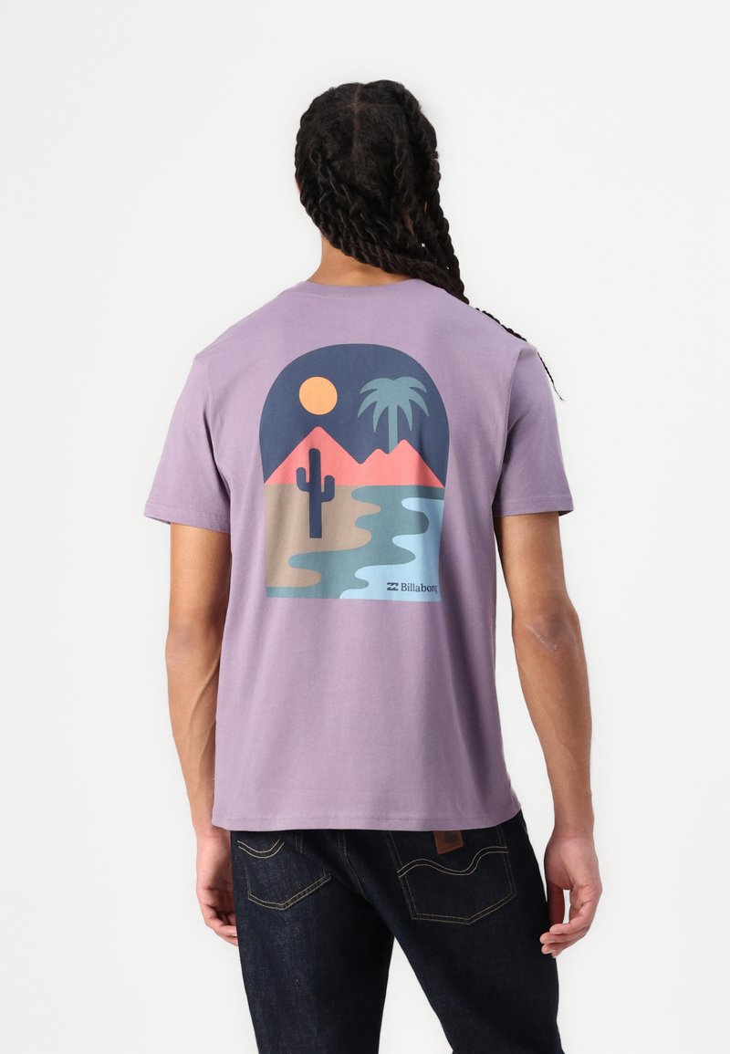 T-shirt en coton violet avec un motif dans le dos représentant un paysage désertique avec des montagnes, un cactus, une rivière, un soleil et un palmier.