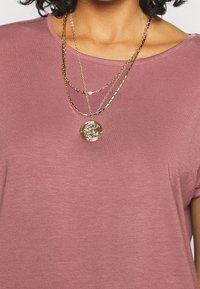 Lichtroze shirt met korte mouwen en een ronde halslijn. Twee gouden gelaagde kettingen met een ronde hanger met ingewikkelde details.