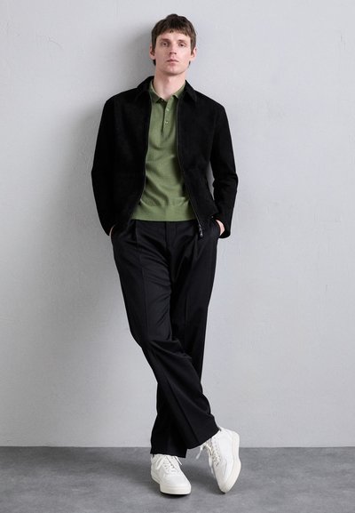 Veste zippée en daim noir sur un polo vert olive, associée à un pantalon noir et des baskets blanches. Design simple mais moderne.