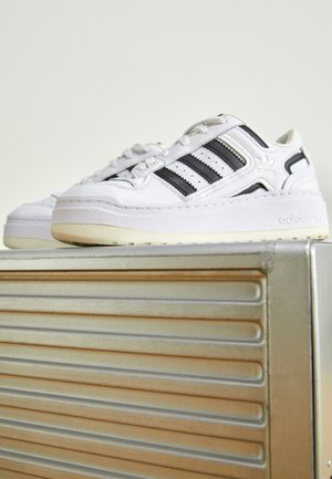 Hvide Adidas sneakers med sorte striber, hvilende på en metalsurface med horisontale riller, fotograferet mod en ensartet lys baggrund.