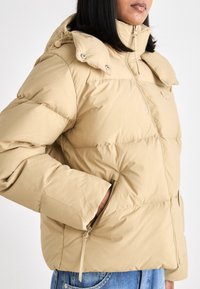 Giacca puffer beige con chiusura a zip, cappuccio staccabile e tasche laterali. Presenta un design trapuntato e una texture liscia.