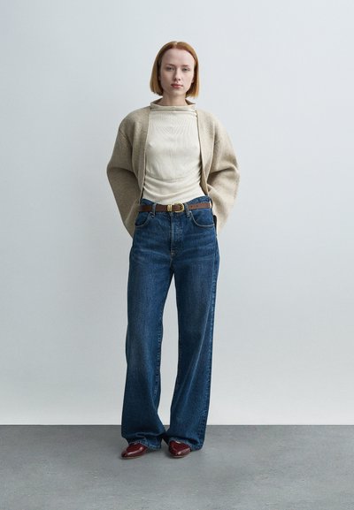 Μπεζ πουλόβερ σε μεγάλο μέγεθος πάνω από αμάνικο κρεμ τοπ με υφή, τζιν παντελόνι ψηλόμεσο σε μπλε απόχρωση και καφέ δερμάτινη ζώνη με χρυσή αγκράφα. Καφέ παπούτσια.