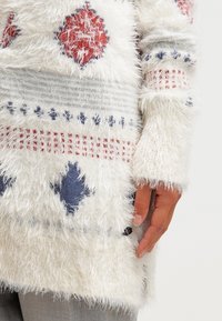 Bílý chlupatý svetr s červenými a modrými geometrickými vzory, s jemnou texturou, dlouhými rukávy a střední délkou, s viditelnou rukou, která ho drží.