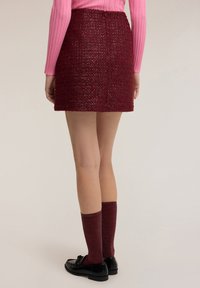 Bordeauxrode tweed minirok met een gestructureerd oppervlak, gecombineerd met kniekousen en zwarte loafers. Gevangen vanuit de achterkant.