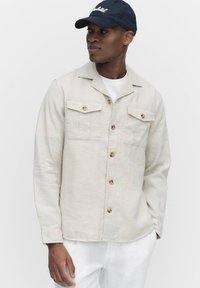 Lichtbeige linnen jasje met een knoopsluiting aan de voorkant, twee borstzakken en lange mouwen. Gecombineerd met een wit shirt en een marineblauwe pet.