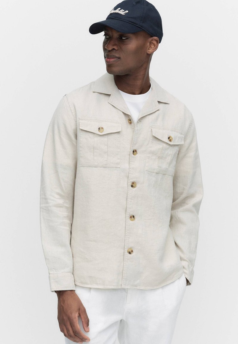 Lichtbeige linnen jasje met een knoopsluiting aan de voorkant, twee borstzakken en lange mouwen. Gecombineerd met een wit shirt en een marineblauwe pet.