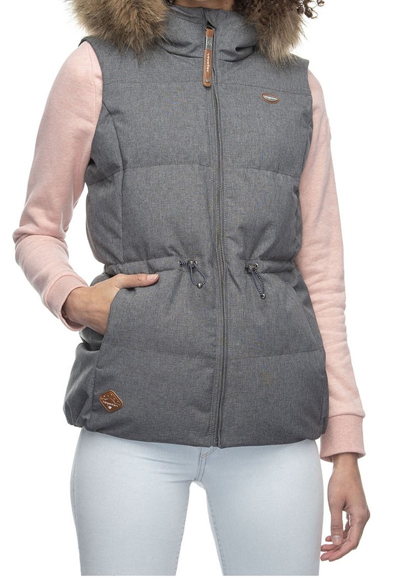Femme portant une veste matelassée grise sans manches avec capuche bordée de fourrure par-dessus un haut à manches longues rose et un jean bleu clair, main dans la poche de la veste.