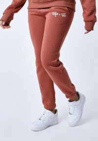 Pantalones de chándal de color marrón rosado con puños elásticos y un logo blanco, combinados con zapatillas blancas. Tela suave con un diseño casual.