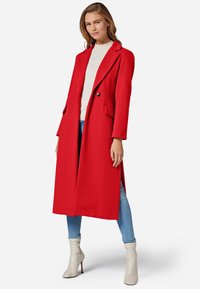 Manteau long rouge en tissu texturé, doté d'un col à revers, d'une fermeture à un bouton et de poches latérales. Porté par-dessus un haut beige clair.
