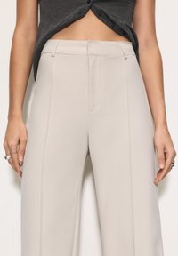 Pantalons larges beiges en tissu lisse avec des plis sur le devant et des poches sur les côtés ; associés à un haut gris foncé, laissant entrevoir le ventre.