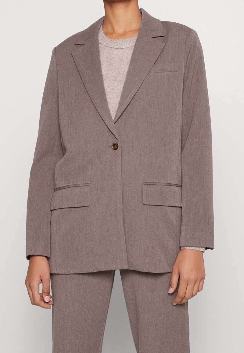 Blazer oversize taupe avec fermeture par un seul bouton, porté sur un pull en maille beige clair, associé à un pantalon taupe assorti.