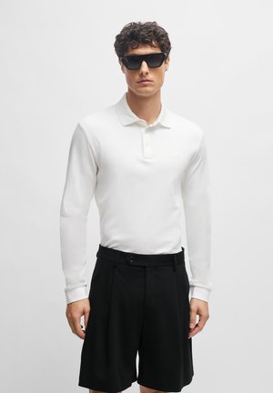 White long-sleeve polo shirt with a collar, button placket, paired with black pleated shorts. Model wears black sunglasses.