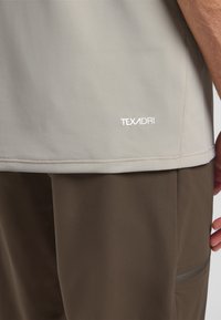 Lichtgrijze shirt met korte mouwen en een textuurafwerking, met een wit logo "TEXADRI" nabij de zoom, gedragen met een bruine broek.