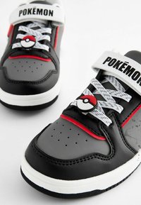 Schwarze und graue Sneaker verfügen über einen weißen Riemen mit der Aufschrift "POKEMON", rote Akzente und ein Poké Ball-Emblem im Schnürbereich. Strukturierte Materialien und sichtbare Belüftungsöffnungen.