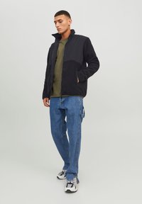 Svart fleecejakke med teksturerte paneler, grønn T-skjorte, blå denimjeans og svart/hvite sneakers, stående mot en lys bakgrunn.