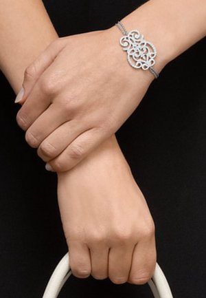 Hands crossed at wrists, wearing a delicate silver bracelet with intricate swirls, holding a white bag handle against a black background.