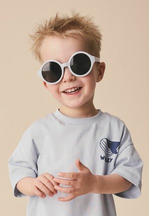 Młody chłopiec z nastroszonymi włosami, noszący duże okrągłe białe okulary przeciwsłoneczne i jasnoniebieską koszulkę z grafiką wieloryba, uśmiechający się na beżowym tle.