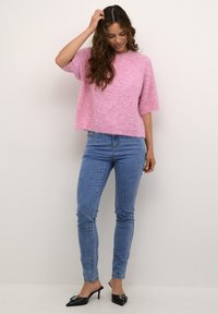 Maglione knit rosa chiaro e peloso con maniche corte; abbinato a jeans in denim blu aderenti e mule neri con tacchi e decorazioni.