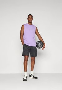 Vijolična brezrokavnik majica, črne mrežaste kratke hlače, drži košarkarsko žogo. Črno-bele superge s teksturirano podplatom. Enostavna oblika, športni kroj.