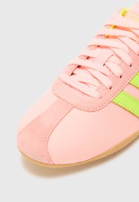 Gros plan d'une basket rose avec un détail en daim, des lacets rose clair et des rayures vert vif sur une semelle claire sur un fond uni.
