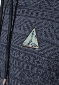 Dunkelblauer Pullover mit strukturiertem Strickmuster, featuring einem dreieckigen Aufnäher, der einen Leuchtturm und Text in einer kontrastierenden Farbe zeigt.